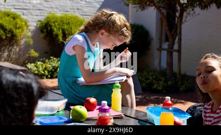 Das kaukasische Mädchen konzentriert sich auf ihre Schularbeit im Freien, umgeben von Gleichaltrigen Stockfoto