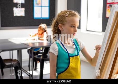 Weißes Mädchen mit blonden Haarfarben auf einer Staffelei, mit blauer Schürze in der Schule Stockfoto