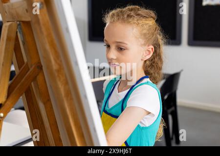 Weißes Mädchen mit lockigen blonden Haaren malt in der Schule auf einer Staffelei Stockfoto