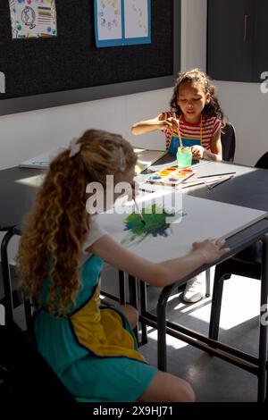 Zwei Mädchen, eine Kaukasierin und eine Birasie, malen am Schultisch, Kopierraum Stockfoto