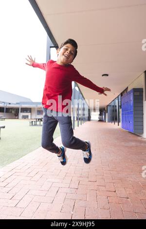 In der Schule im Freien springt ein birassischer Junge, der ein rotes Hemd trägt, vor Freude Stockfoto