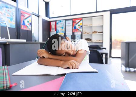 Erschöpfter birassischer Junge ruht während des Studiums in einem hellen Klassenzimmer auf dem Schreibtisch. Stockfoto