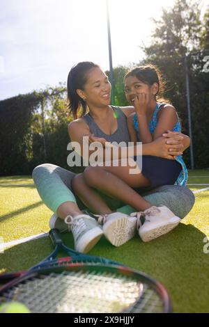 Im Freien sitzen Mutter und Tochter auf dem Tennisplatz, lächeln und umarmen Stockfoto