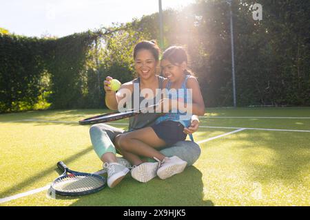 Zweirassige Mutter und Tochter lächeln, spielen draußen mit ihren Schlägern Tennis Stockfoto