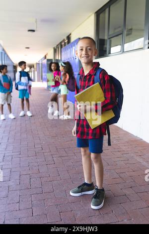 Ein birassischer Junge mit Rucksack und gelbem Notizbuch steht lächelnd in einem Schulflur. Andere Kinder interagieren im Hintergrund, was auf a b hindeutet Stockfoto