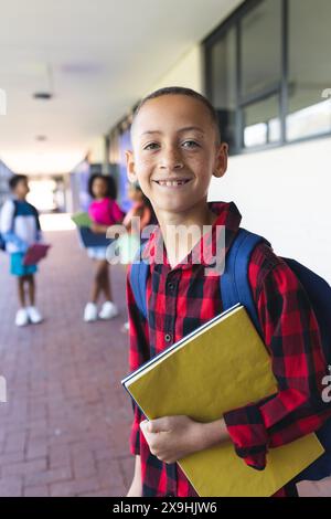 Ein birassischer Junge mit Rucksack und gelbem Notizbuch lächelt in der Schule Stockfoto