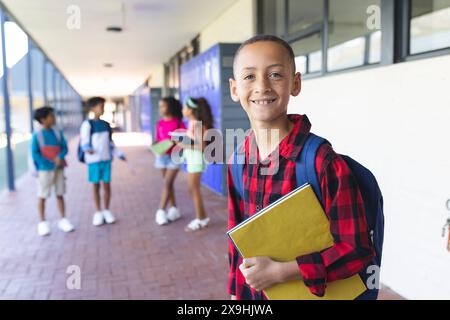 Ein birassischer Junge mit Rucksack steht lächelnd und hält in der Schule eine gelbe Mappe Stockfoto