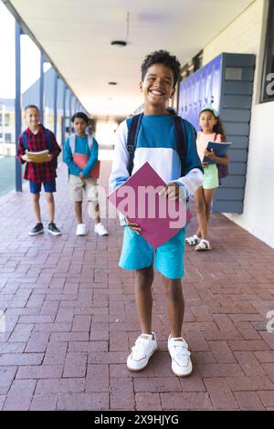 Ein birassischer Junge im Vordergrund lächelt, hält einen Ordner, mit Klassenkameraden hinter der Schule Stockfoto