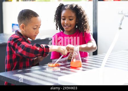 Junge und Mädchen mit birassischer Rasse nehmen in der Schule an einem wissenschaftlichen Experiment Teil, in lässiger Kleidung. Sie konzentrieren sich auf das Mischen von Substanzen in Laborumgebungen, Stockfoto