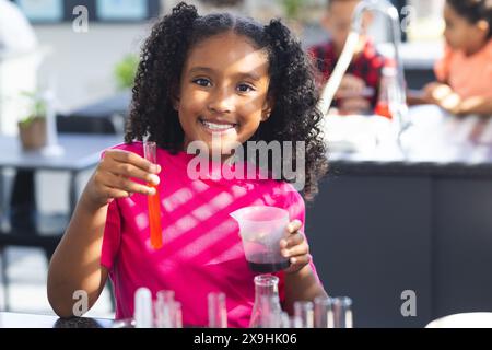 Ein birassisches Mädchen mit lockigen Haaren lächelt, während er eine Pipette und einen Becher in einem Wissenschaftskurs hält Stockfoto