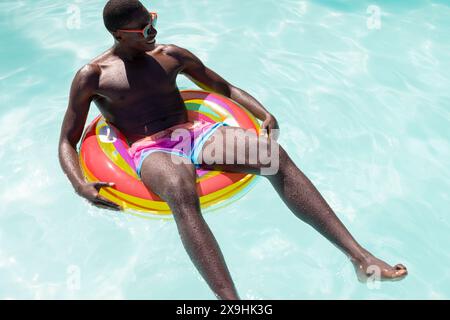 Ein junger Afroamerikaner, der draußen auf einem farbenfrohen Wagen liegt. Sonnenbrille und pinkfarbene Shorts tragen, Sonnenschein in entspannter Poolatmosphäre genießen, Stockfoto