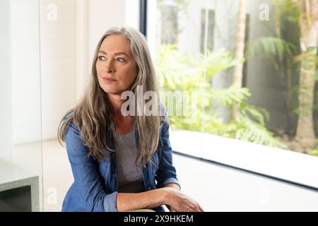 Zu Hause sitzt eine ältere kaukasische Frau mit langen grauen Haaren und blauen Augen drinnen. Trage ein blaues Denim-Hemd über grauem Oberteil und schaue nachdenklich aus Stockfoto