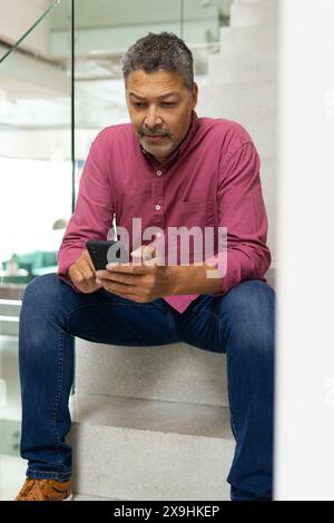 Zu Hause sitzt ein zweirassiger älterer Mann mit grauen Haaren und Bart auf der Treppe mit dem Telefon. Mit einem pinkfarbenen Hemd, einer blauen Jeans und braunen Schuhen Stockfoto