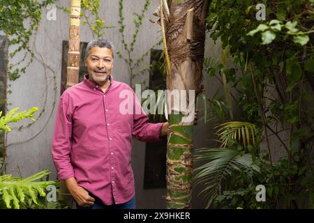 Draußen, zweirassige ältere Männer, die im üppigen Garten stehen und Baumstamm berühren. Er hat kurze, lockige graue Haare, einen Bart und trägt einen kastanienbraunen Button-up Stockfoto