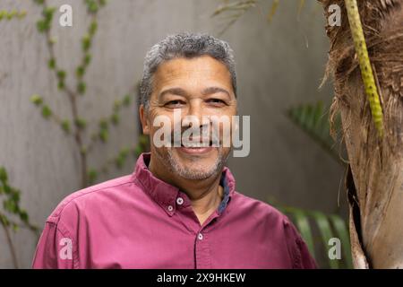 Zweirassiger älterer Mann mit kurzen grauen Haaren und Ziegenbart, der draußen lächelt. Er trägt ein kastanienbraunes Hemd und steht neben einem Baum mit grüner lea Stockfoto