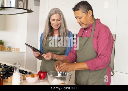 Zu Hause backen verschiedene Seniorenpaare in der Küche, kaukasische Frau mit Tablette. Zweirassiger Ehemann, der Eier in eine Schüssel knackt, beide mittleren Alters, tragen eine Stockfoto