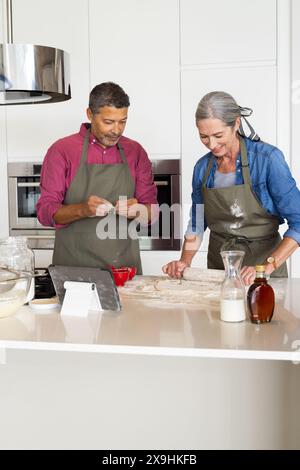 Zu Hause backen verschiedene Seniorenpaare zusammen in moderner Küche. Birassischer Mann mit kurzen lockigen Haaren und kaukasische Frau mit langen grauen Haaren, beide wea Stockfoto