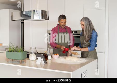 Zu Hause backen diverse Seniorenpaare in moderner Küche, beide tragen Schürzen. Reife weiße Frau mit langen grauen Haaren, die Teig Rollen, reife Biracia Stockfoto