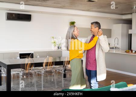 Zu Hause tanzt ein vielseitiges Seniorenpaar zusammen in einer modernen Küche, lächelt, Kopierraum. Birassischer Ehemann mit kurzen grauen Haaren und kaukasische Ehefrau mit Stockfoto