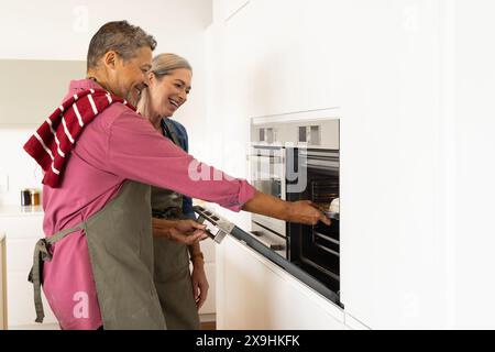 Zu Hause backen verschiedene Seniorenpaare in moderner Küche mit Schürzen zusammen. Kaukasische Seniorin mit grauen Haaren lächelt während Afroamerikaner Stockfoto