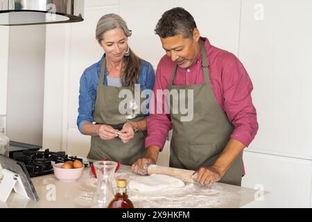 Zu Hause, verschiedene Seniorenpaare in der Küche, die zusammen Teig machen, beide tragen Schürzen. Kaukasische Frau mit langen grauen Haaren und birassischer Mann mit kurzem Stockfoto