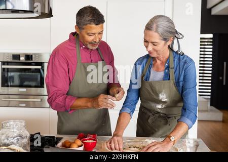 Zu Hause backen verschiedene Seniorenpaare zusammen in der Küche, beide lächeln in Schürzen. Birassischer Mann mit kurzen grauen Haaren und Bart und kaukasische Frau wi Stockfoto