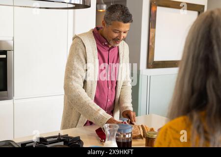 Zu Hause ein vielseitiges Seniorenpaar in moderner Küche; ein birassischer Mann mittleren Alters schneidet Brot. Reife kaukasische Frau mit langen grauen Haaren in der Nähe, W Stockfoto