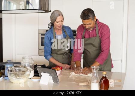Zu Hause backen verschiedene Seniorenpaare in der modernen Küche zusammen, beide in Schürzen. Kaukasische Frau mit grauem Haar und birassischer Mann mit kurzen dunklen Haaren Stockfoto