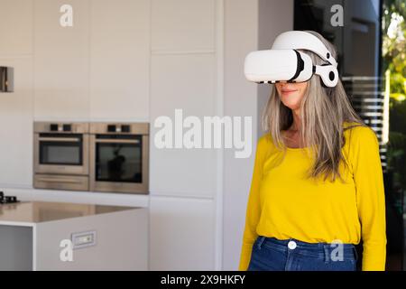 Zu Hause trägt eine kaukasische Seniorin ein Virtual-Reality-Headset in einer modernen Küche, Kopierraum. Lächelnd und mit gelbem Hemd und Jeans Stockfoto