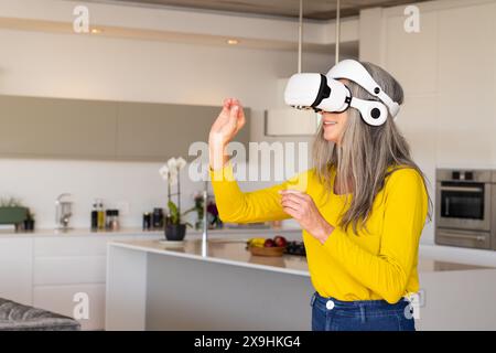 Zu Hause, kaukasische Seniorin in der modernen Küche mit VR-Headset, lächelnd, Kopierraum. Mit gelbem Hemd und Jeans und virtuellem umfeld Stockfoto