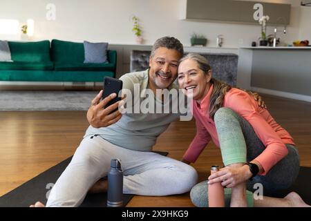 Zu Hause lächelt ein vielseitiges Seniorenpaar für Selfie auf Yogamatten im Wohnzimmer. Birassischer Ehemann mit kurzen lockigen Haaren und grauem Bart, kaukasische Ehefrau wi Stockfoto
