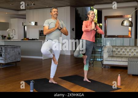 Verschiedene Seniorenpaare üben Yoga zu Hause auf Matten in ihrem Wohnzimmer. Kaukasierin mittleren Alters mit grauem Haar und birassischer Mann mittleren Alters BA Stockfoto
