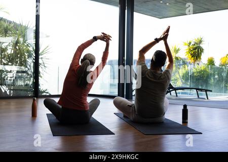 Verschiedene Seniorenpaare sitzen auf Yogamatten in einem hellen Zimmer mit großen Fenstern zu Hause. Die Arme über den Kopf Strecken, von der Kamera weg Stockfoto