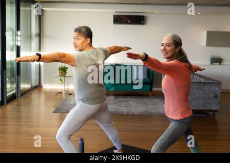 Zu Hause praktizieren verschiedene Seniorenpaare gemeinsam Yoga in einem hellen Wohnzimmer. Birassischer Mann und kaukasische Frau, beide reif, dehnend in Krieger po Stockfoto
