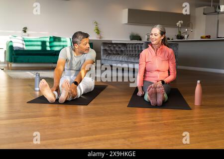 Zu Hause ein vielseitiges Seniorenpaar, das sich auf Yogamatten ausdehnt und sich gegenseitig anlächelt. Birassischer Mann mit kurzen grauen Haaren und Bart, kaukasische Frau mit langem Stockfoto