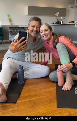 Zu Hause sitzen verschiedene Seniorenpaare auf Yogamatten im Wohnzimmer und machen Selfie. Birassischer Ehemann mit kurzen grauen Haaren und Bart, kaukasische Ehefrau mit Weib Stockfoto