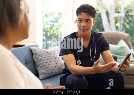 Zu Hause erklärt die birassische männliche Krankenschwester der älteren Patientin aufmerksam. Sie befinden sich in einem gemütlichen Wohnzimmer mit einem großen Fenster, das einen üppigen Garten zeigt Stockfoto