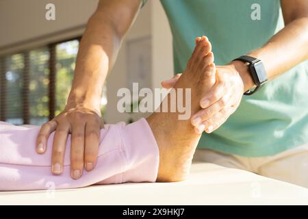 Zu Hause, birassischer männlicher Physiotherapeut, der den Fuß einer älteren Patientin behandelt. Sonnenlicht strömt durch große Fenster in einer hellen, modernen Klinik mit W Stockfoto