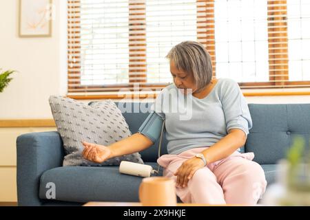 Zu Hause, zweirassige Seniorin, die den Blutdruck mit dem Monitor überprüft. Auf einer blauen Couch in einem hellen, gemütlichen Wohnzimmer mit großen Fenstern und Holz Stockfoto