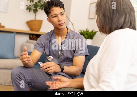 Zu Hause berät eine junge, birassische Krankenschwester eine ältere Patientin. Sie sind in einem gemütlichen, gut beleuchteten Wohnzimmer mit Pflanzen und komfortablen Möbeln, u Stockfoto