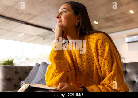 Zu Hause entspannte junge kaukasische Frau, hielt Buch und sah nachdenklich aus. Sie sitzt auf einer bequemen grauen Couch in einem modernen, gut beleuchteten Wohnbereich Stockfoto