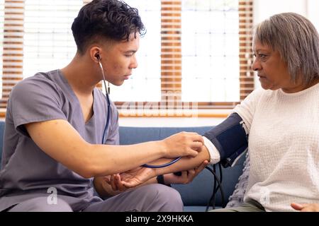 Zu Hause nimmt eine birassische männliche Krankenschwester den Blutdruck einer älteren Patientin. Sie befinden sich in einem hellen, gut beleuchteten Zimmer mit großen Fenstern und komfortablen Zimmern Stockfoto