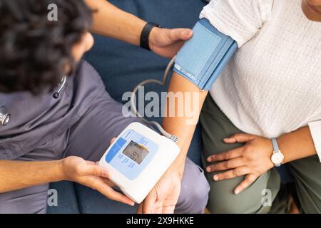 Zu Hause kontrolliert eine junge, birassische Krankenschwester den Blutdruck einer älteren Frau. Sie sind in einem medizinischen Büro mit blauem Hintergrund und medizinischer Ausrüstung Stockfoto
