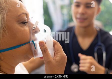 Eine birassische Krankenschwester hilft einer älteren Patientin mit einer Atemschutzmaske zu Hause. Sie befinden sich in einer hellen, gut beleuchteten Klinik, in der medizinische Geräte sichtbar sind Stockfoto