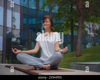 Entlasten, Ideen abwickeln. Friedliche freischaffende Mitarbeiterin, die Yoga-Übungen praktiziert, während sie nach einem harten Arbeitstag im Büro in einem Haufen unter Glashochhäusern sitzt und die Hände in Mudra hält Stockfoto