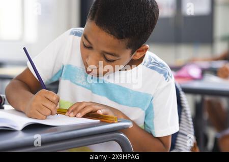 Ein birassischer Junge schreibt in Notizbuch, trägt ein gestreiftes Hemd Stockfoto