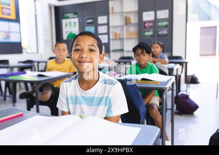 Ein birassischer Junge mit einem weißen T-Shirt lächelt in einem Klassenzimmer, Bücher öffnen sich auf den Schreibtischen Stockfoto