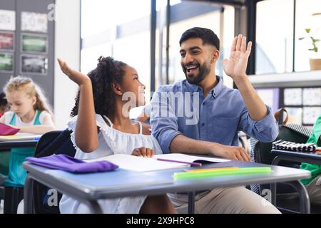 Junger asiatischer männlicher Lehrer und birassisches Mädchen High-Five in einem Klassenzimmer Setting Stockfoto