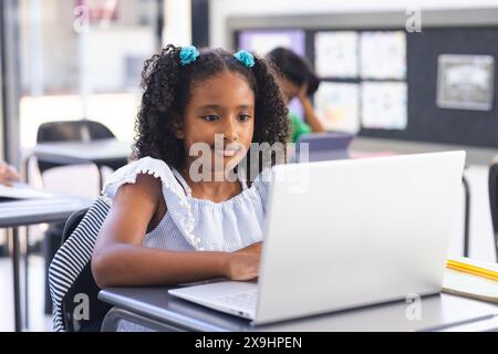 In der Schule, im Klassenzimmer, ein junges birassisches Mädchen mit lockigem Haar, das sich auf den Laptop konzentriert. Sie trägt eine weiße Bluse, ihre Klassenkameradin im Hintergrund-Concentrati Stockfoto