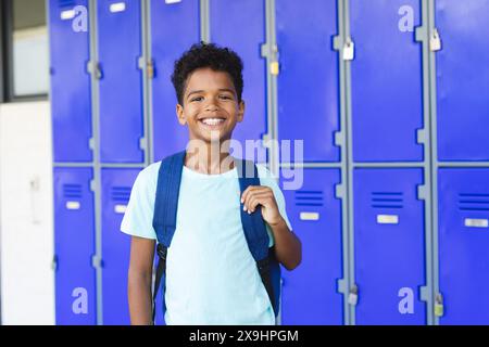 Ein birassischer Junge mit Rucksack steht lächelnd vor blauen Schließfächern in der Schule Stockfoto